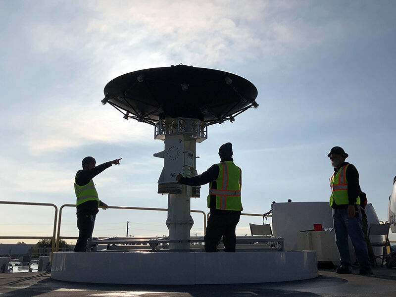 Megalodon dimensionally tunable reflector is deployed in the field. Hardware is on a rooftop, and there is a blue sky and bright sun above. Three technicians are working around the structure. The reflector dish is attached to a large column and is directed up at the sky, with electronics equipment and wiring visible. 